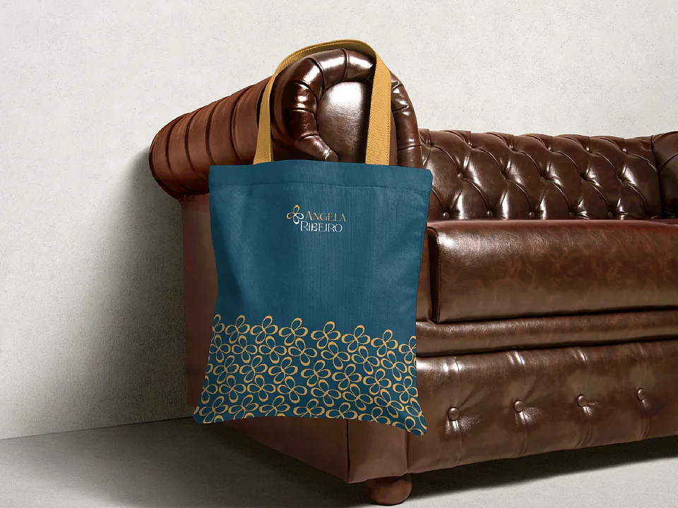 Tote bag azul petróleo com barra de pétalas douradas, apoiada em sofá de couro