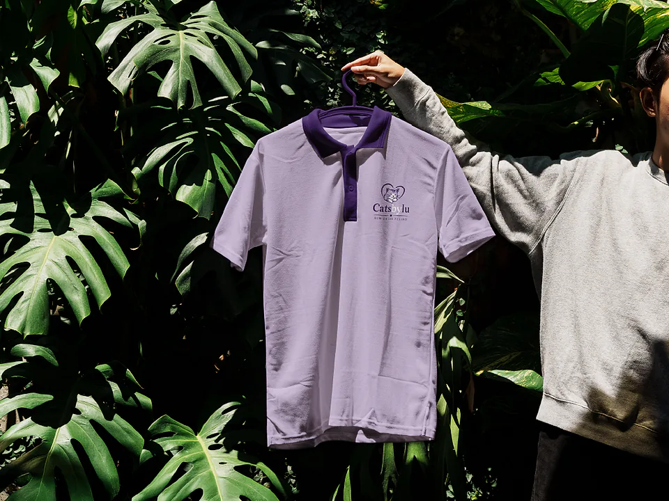 Camisa polo lilás com a marca Catsbylu bordada, pendurada entre folhagens
