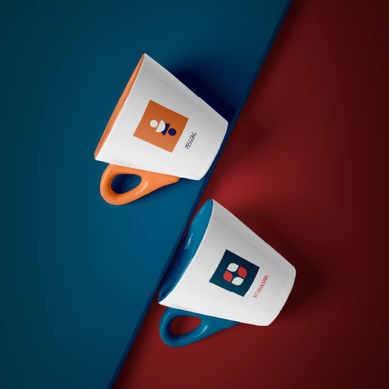Duas canecas brancas, uma com alça laranja e outra com alça azul, cada uma com um símbolo do evento, sobre fundos vermelho e azul